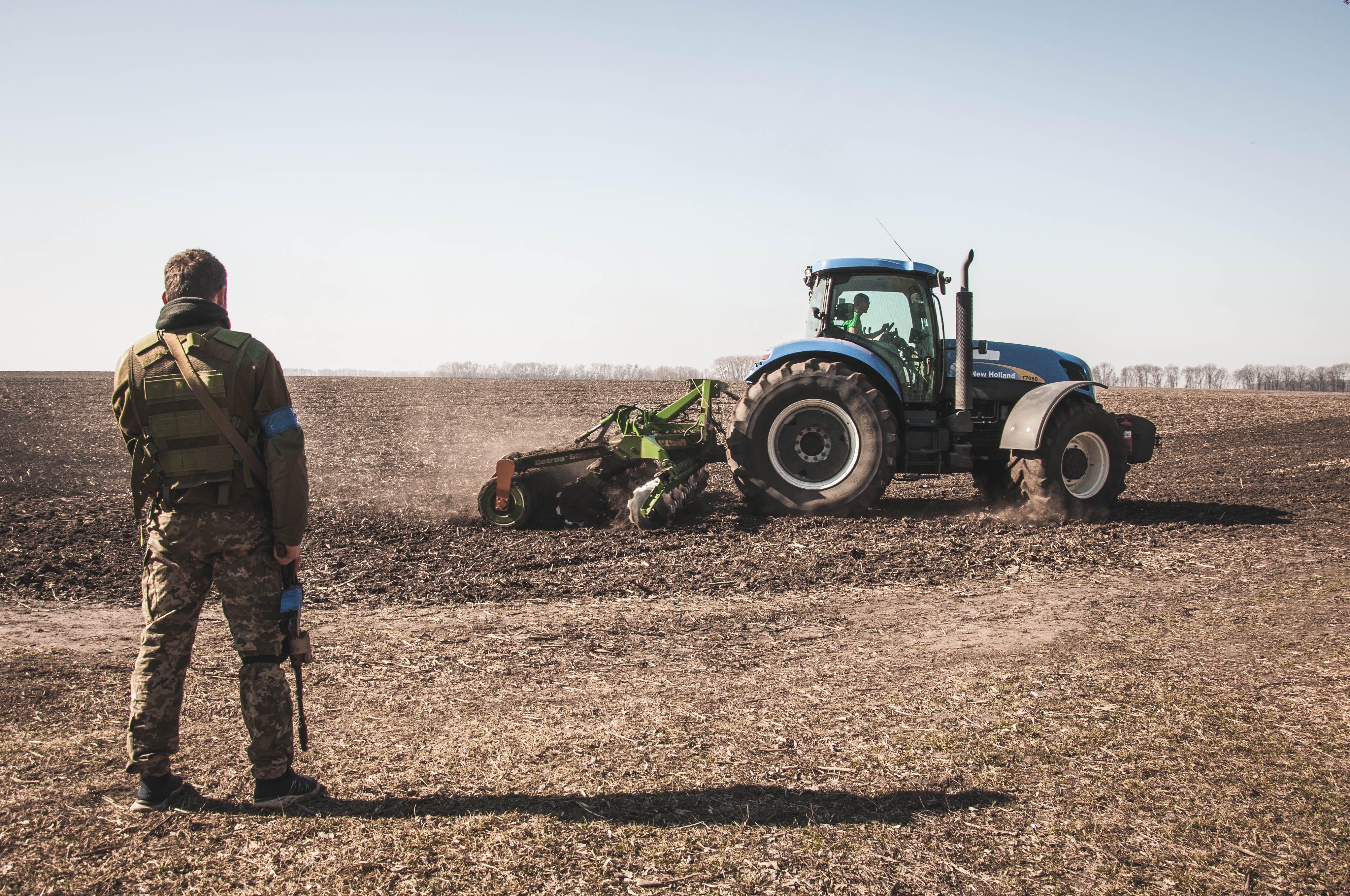 Ein ukrainischer Soldat betrachtet einen Traktor, der ein Feld während des Kriegs in der Ukraine pflügt Ein ukrainischer Soldat betrachtet einen Traktor, der ein Feld während des Kriegs in der Ukraine pflügt