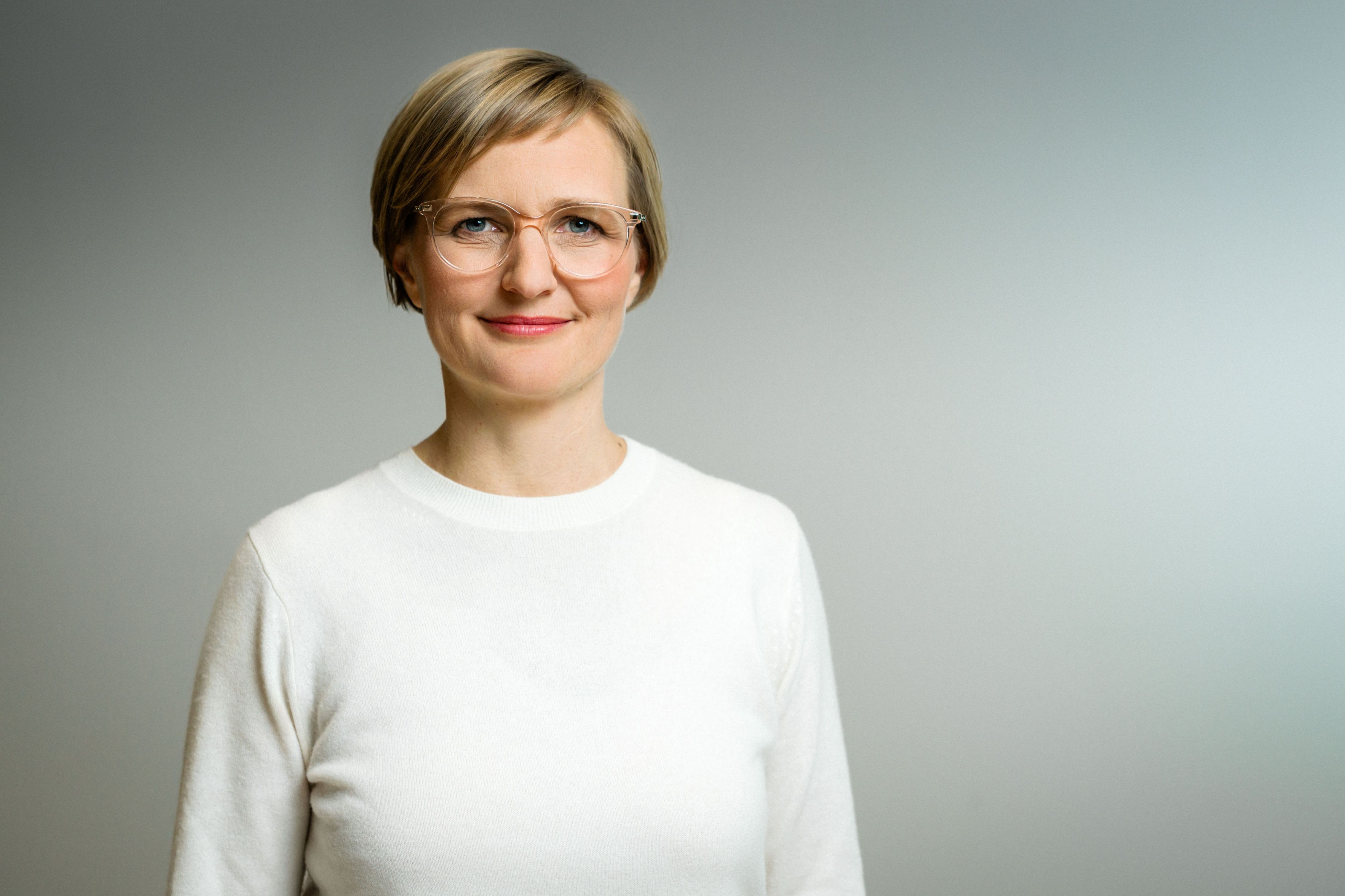 Person mit kurzem Haar und Brille steht vor grauem Hintergrund und lächelt leicht