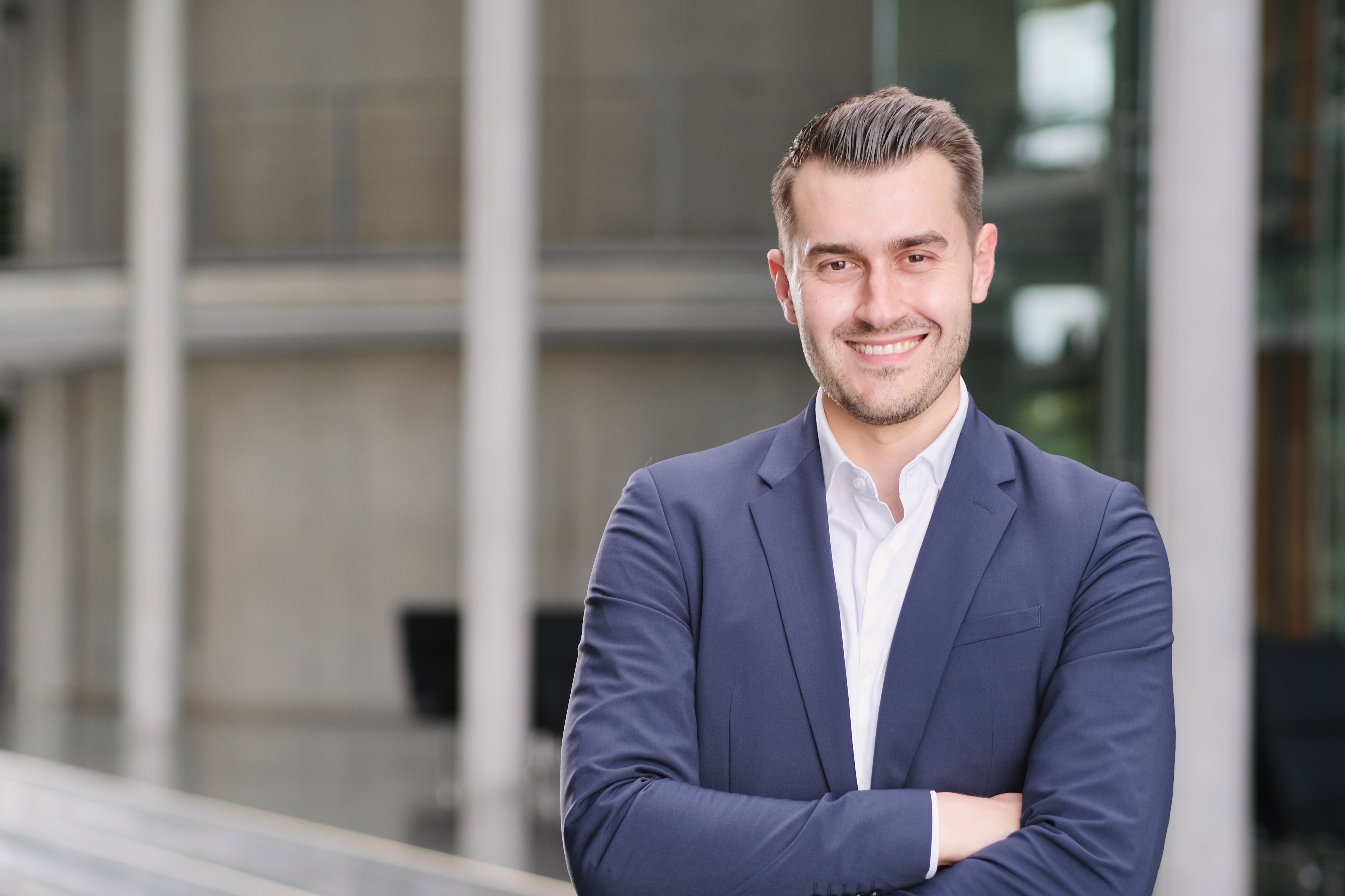Person mit kurz gescheiteltem Haar steht mit verschränkten Armen vor moderner Betonfassade