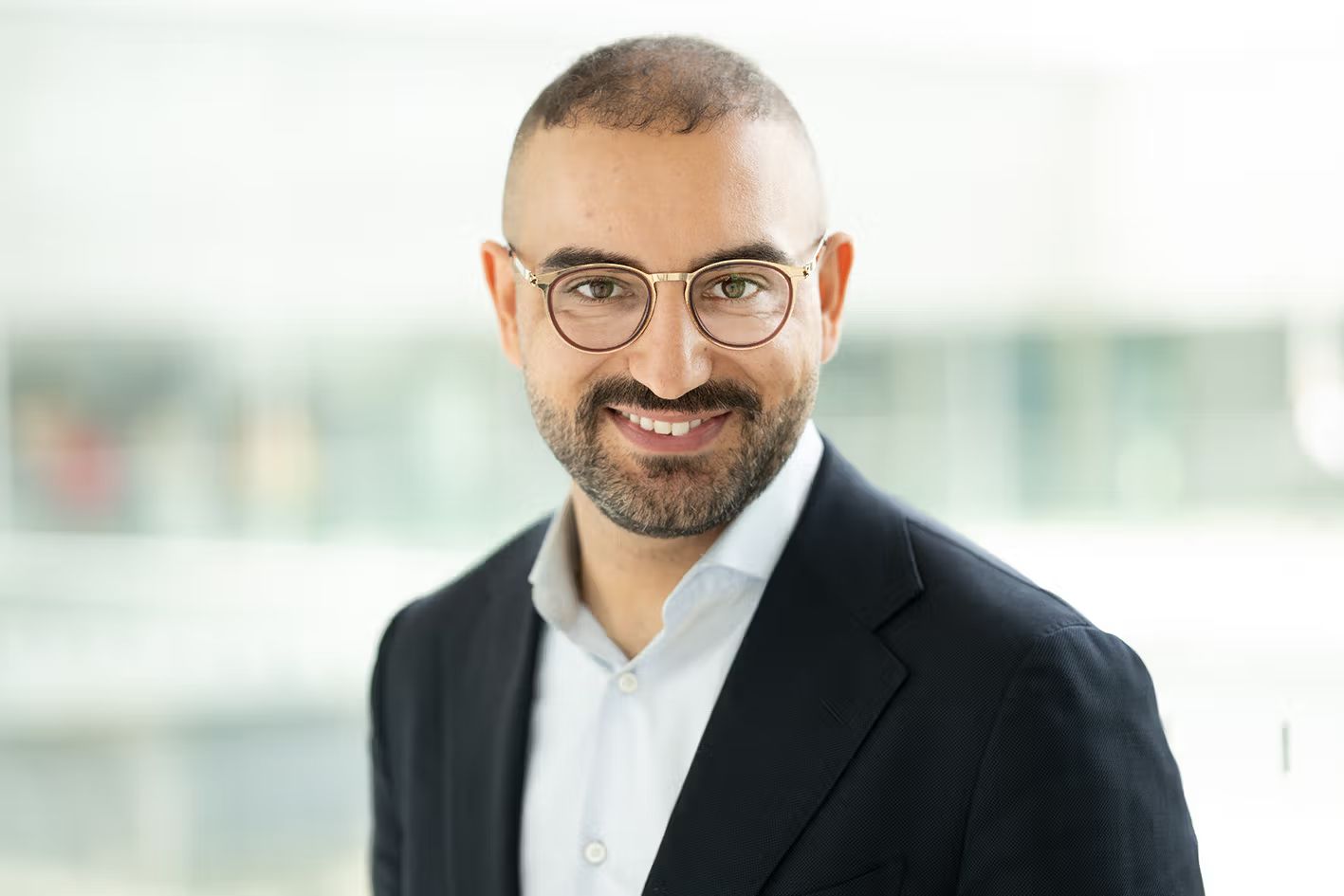 Person mit kurzem Haar, kurzem Bart und runder Brille steht vor heller Glasfassade und lächelt in die Kamera.