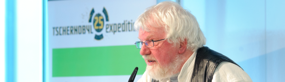 Mann mit grauen Haaren und Lederweste an Mikrofon vor Schild "Tschernobyl 25 - expeditionen"