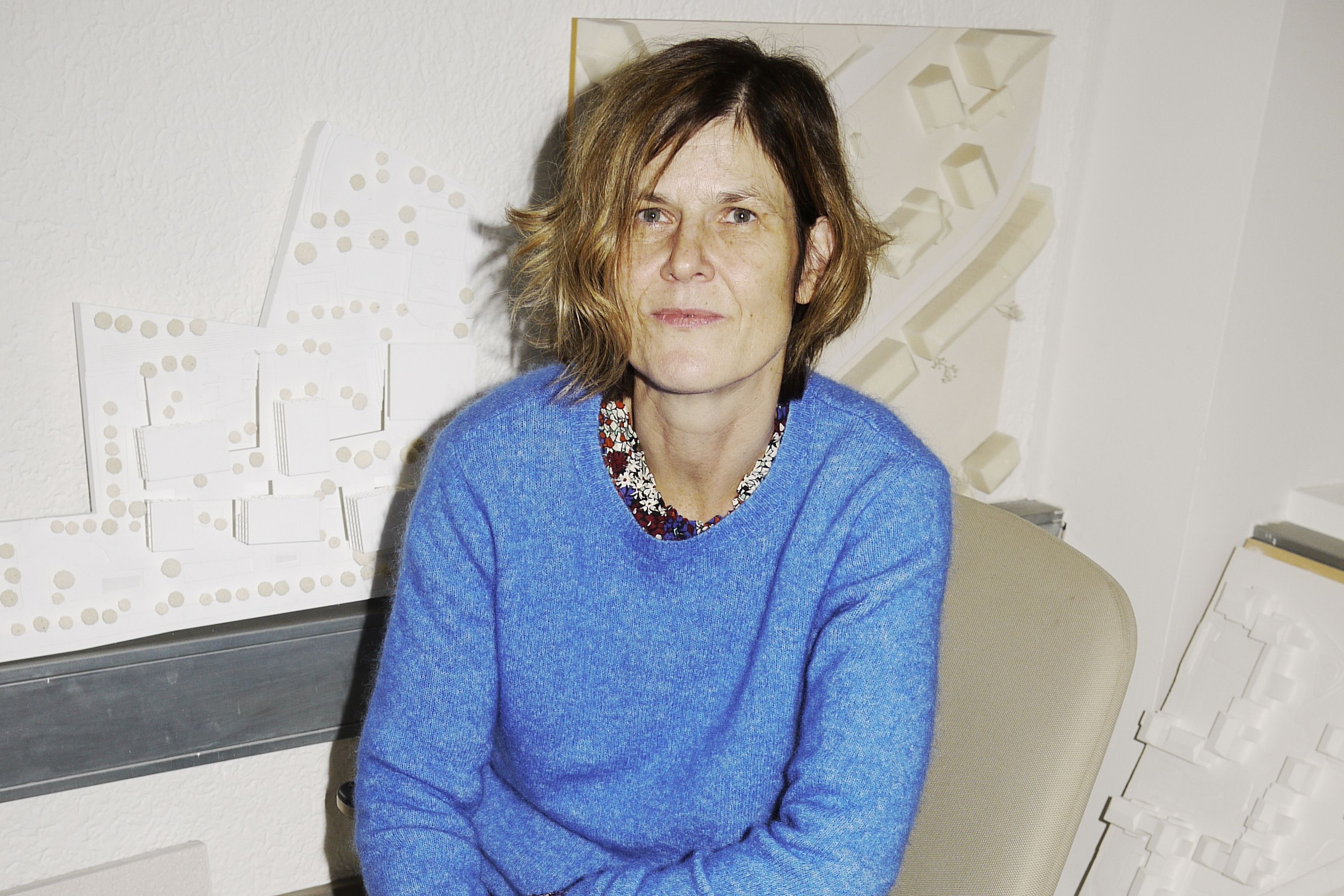 Person sitzt auf Stuhl vor Wand mit Architekturmodellen, Blick zur Kamera, kurze Haare, ein Modell leicht hinter dem Kopf.