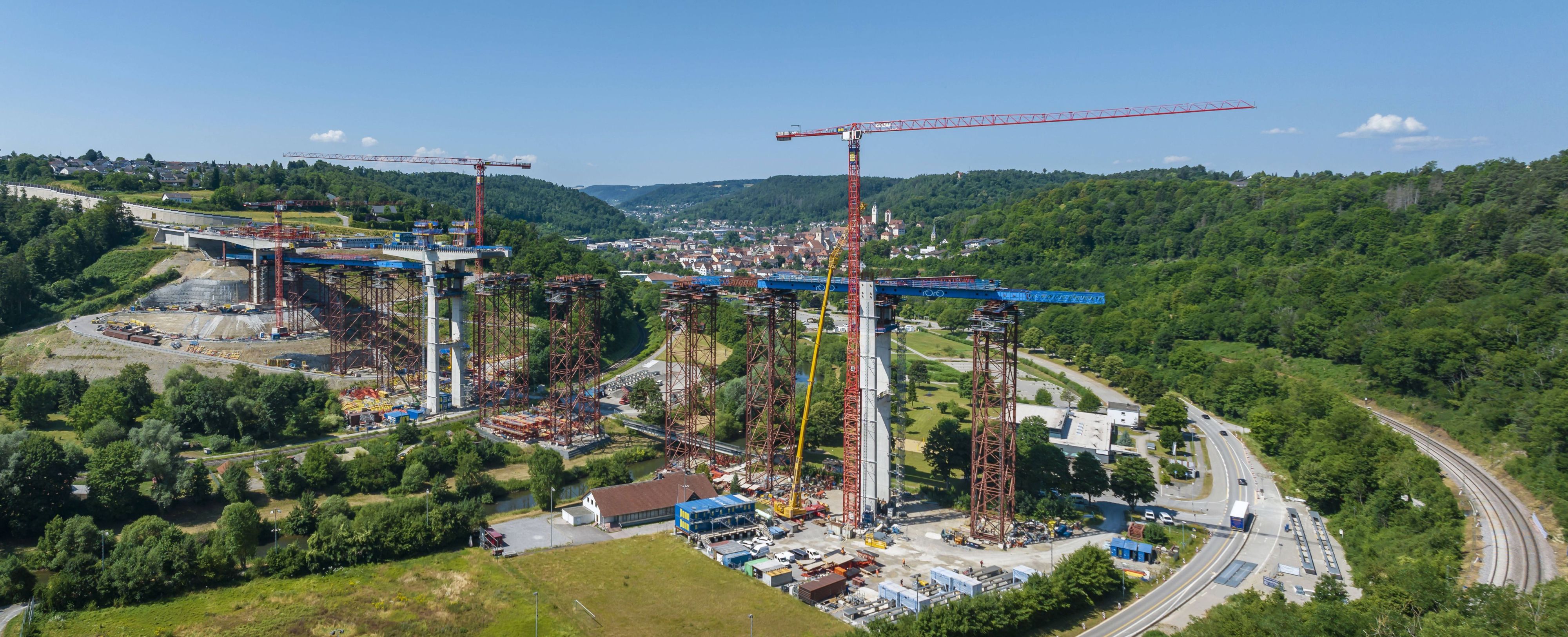 Große Brückenbaustelle im Tal, mehrere Kräne und Pfeiler, Straße und Bahn rechts, Ortschaft im Hintergrund, viel Grün