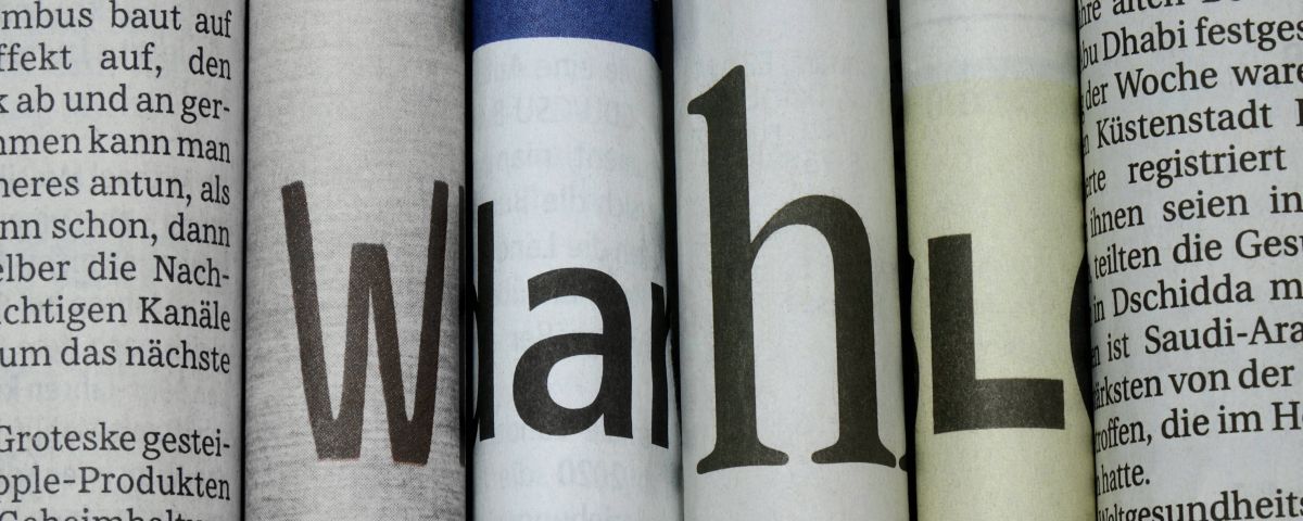 Mehrere eng nebeneinander aufgerollte Zeitungsseiten mit großen Buchstaben und angeschnittenen deutschen Texten.