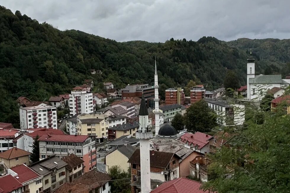 Blick über eine Kleinstadt mit Moscheen und einer Kirche, umgeben von bewaldeten Hügeln unter dicht bewölktem Himmel.
