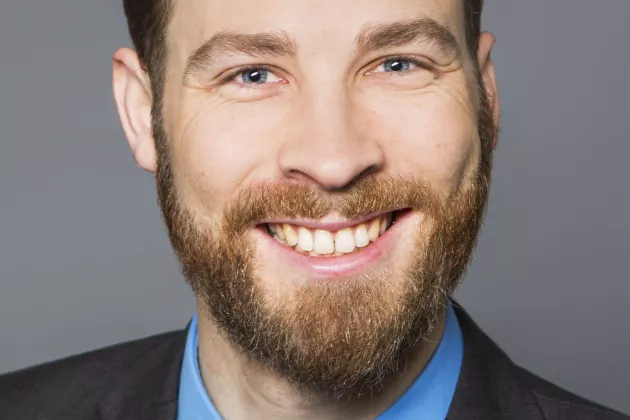 Ein junger Man mit Bart und Anzug schaut breit lächelnd in die Kamera