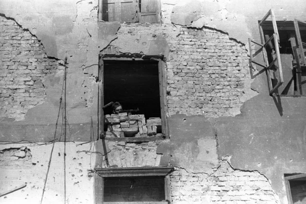Foto (schwarz-weiß): Ein bewaffneter Mann zielt aus einem teils zerstörten Fenster, hinter einer Mauer aus Ziegeln. Die Fassade ist stark beschädigt.