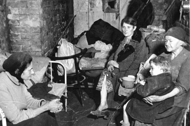 Foto (schwarz-weiß): Vier Frauen, darunter eine mit einem Kind auf dem Schoß, sitzen in einem Kellerraum, umgeben von Decken und Habseligkeiten.