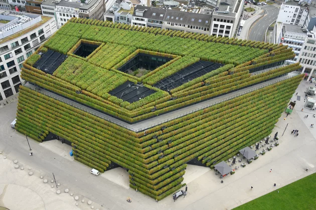 Begrüntes Gebäude mit Terrassenpflanzung. Dach mit offenen Innenhöfen, umgeben von städtischer Bebauung und Gehwegen.