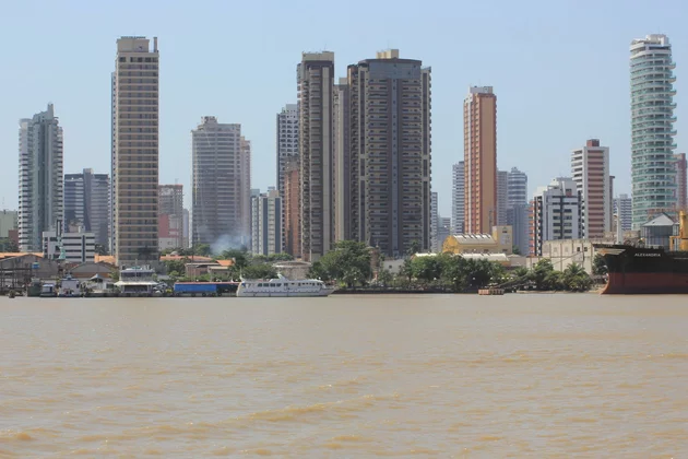 Mehrere Hochhäuser hinter Flussufer mit Boot und Häusern im Vordergrund, Rauch steigt zwischen Bäumen auf.