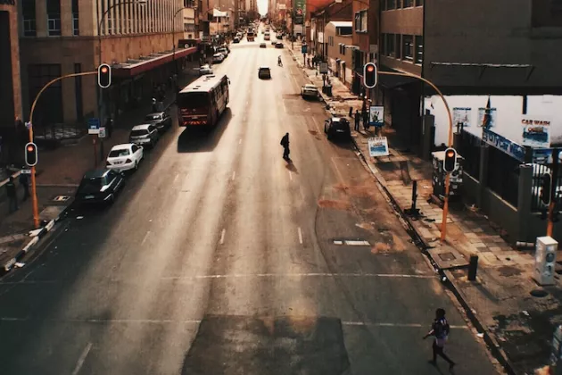 Foto: Breite Stadtstraße in einem Geschäftsviertel bei Sonnenuntergang. Ein Bus und Autos fahren, zwei Fußgänger*innen überqueren die Straße. Hochhäuser im Hintergrund.