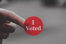 One hand is holding a sticker with "I Voted" written on it against a blurred background.