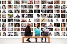 Drei Menschen sitzen auf einer Bank vor einer großen Foto-Wand mit vielen unterschiedlichen Gesichtern.