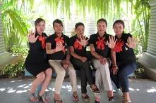 Five women in black dresses are sitting on a bench and showing their show their outstretched hands 