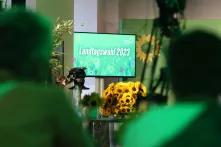 Ein großer Bildschirm zeigt eine grüne Wiese und darauf den Schriftzug "Landtagwahl 2023", davor liegen ein großer Strauß Sonnenblummen, im Hintergrund stehen Aufsteller der Partei Bündnis 90/Die Grünen. Im Vordergrund sind schemenhaft Personen mit Kameras zu erkennen.