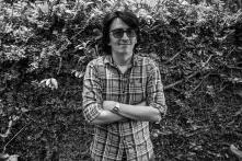 Portrait of Bill Barreto. Black and white photo. He is wearing sunglasses, has his arms crossed, and is standing in front of a hedge.
