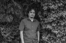 Portrait of Oswaldo Hernández. Black and white image. He is standing among trees.