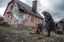 Eine Person steht mir einem Hund vor einem Einfamilienwohnhaus mit rosa Fassade. An der Vorderseite ist ein großer Teil der Fasssade weggesprengt und knapp unter dem Dach ist ein großes Loch zu sehen.