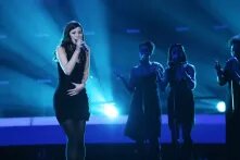 Lena Meyer-Landrut steht auf einer blau ausgeleuchteten Bühne und hält ein Mikrofon in der Hand. Dahinter, weniger hell beleuchtet, drei Sängerinnen. Alle vier tragen schwarze Kleider.