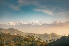 Eine bewaldete Hügelkette mit einigen nepalesischen Häusern, dahinter die schneebedeckten Gipfel des Himalaya unter blauem Himmel.