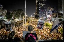 Foto: Nachtaufnahme in Tel Aviv, viele Demonstrant*innen mit Fahnen und Plakaten auf einer Straße, Wolkenkratzer im Hintergrund.