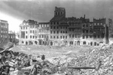 Foto (schwarz-weiß): Eine Reihe zerstörter Gebäude mit ausgebrannten Fenstern. Im Vordergrund liegen Trümmer und Ziegelreste verstreut.