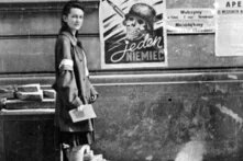 Foto (schwarz-weiß): Eine Frau steht auf einem Gehweg neben einer Wand mit Plakaten. Eines zeigt den Text „Każdy pocisk jeden Niemiec“ ("Jede Kugel ein Deutscher"). Sie trägt eine Armbinde und hält ein Papier.