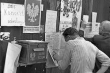 Foto (schwarz-weiß): Zwei Männer lesen eine Wand mit Plakaten. Eines davon zeigt einen weißen Adler, auf einem anderen steht „Żołnierz A.K. nie plotkuj” (Soldat der Heimatarmee, tratsche nicht) 