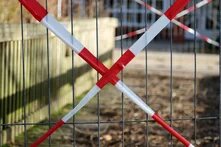 Foto: Ein Metallzaun mit rot-weißem Absperrband, das sich kreuzt. Dahinter ein Weg und ein Holzzaun, im Hintergrund unscharf ein Gebäude.