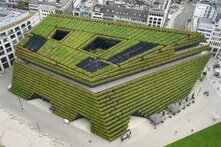Begrüntes Gebäude mit Terrassenpflanzung. Dach mit offenen Innenhöfen, umgeben von städtischer Bebauung und Gehwegen.