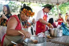 Foto: Zwei Personen mit buntem Kopfschmuck und Schürzen kochen an einem Holztisch im Freien. Weitere Menschen sind im Hintergrund sichtbar.