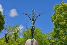 Photo: Statue of a child with outstretched arms holding a crane. Surrounded by trees, blue sky in the background.