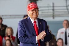 Foto: Donald Trump mit roter „Make America Great Again“-Kappe und Anzug reibt sich grinsend die Hände. Unscharfes Publikum im Hintergrund.
