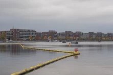Wasserfläche mit gelber Schwimmlinie im Vordergrund, dahinter eine Reihe moderner, brauner Wohngebäude unter grauem Himmel.