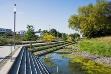 Stufenanlage führt zu einem bepflanzten Wasserbecken, gesäumt von Laternen und modernen Gebäuden im Hintergrund.