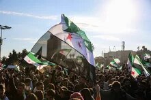 Foto: Menschenmenge bei einer Kundgebung, viele syrische Oppositionsflaggen werden hochgehalten, Sonnenschein im Hintergrund.