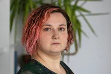 Foto: Person mit lockigem, rosa Haar und Nasenring vor unscharfem Hintergrund mit Pflanze.