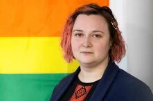 Darya Afanasyeva - Person mit kurzen, rötlichen Haaren steht vor einer Regenbogenflagge. Der Blick ist neutral, der Hintergrund schlicht.