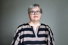 Foto: Person mit grauem Haar und Brille vor neutralem Hintergrund, trägt ein gestreiftes Oberteil.