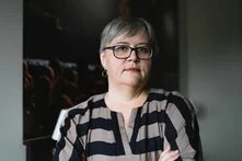 Foto: Person mit grauem Haar und Brille vor dunklem Hintergrund, trägt gestreiftes Oberteil und hat die Arme verschränkt.