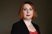 Foto: Person mit kinnlangem, rötlichem Haar vor dunklem Hintergrund, trägt roten Lippenstift und ein Oberteil mit Spitzenbesatz.