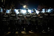 Foto: Eine Reihe von Polizist*innen mit Helmen und Schilden steht nachts eng beieinander, mit grellem Licht im Hintergrund.