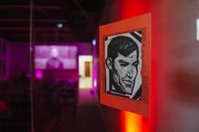 Foto: Nahaufnahme einer Zeichnung eines Gesichts auf rotem Papier, rechts an einer Wand. Unscharf im Hintergrund ein Raum mit violettem Licht.