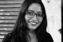 Photo: A smiling person with glasses and long hair stands in front of a closed shutter.