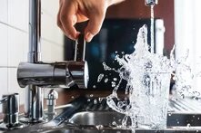 Eine Hand dreht einen Wasserhahn auf, Wasser spritzt in ein überlaufendes Glas in einer Spüle.