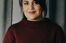 GPT Icon  Foto: Person mit dunklen Haaren im Dutt, trägt einen weinroten Pullover. Hintergrund aus grauen Vorhängen.