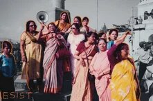 GPT Icon  Photo: A group of women protest on a street. One speaks into a megaphone, others point in different directions. The word "RESIST" is written. The women are highlighted in color, while the background is black and white.