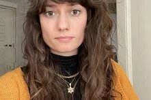 Foto: Person mit langen, lockigen Haaren, Haarspange, schwarzem Oberteil und gelber Strickjacke. Hintergrund zeigt eine weiße Wand und Tür.
