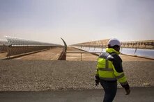 GPT Icon  Foto: Eine Person mit Schutzkleidung und Helm läuft an einer großen Solaranlage mit langen Reihen von Spiegeln in einer Wüstenumgebung entlang.