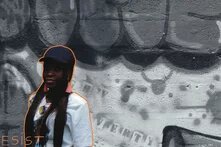 Photo: A woman stands in front of a graffiti-covered wall. She is highlighted in color, while the background is black and white. "RESIST" is written in the bottom left corner.